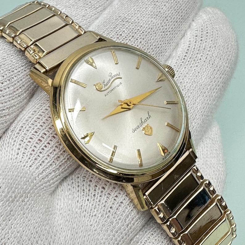 May include: A gold-tone wristwatch with a white face and gold-tone hands. The watch face has the text "Sea Shark" and "Saxon Ricard Automatique" printed on it. The watch is on a gold-tone metal band.