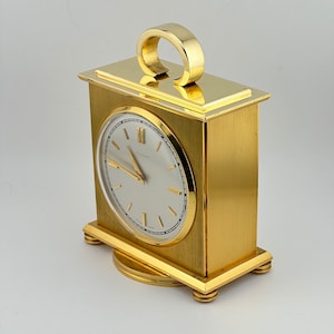 May include: A gold-toned, square-shaped desk clock with a white face and black hands. The clock has a raised, circular handle on the top.