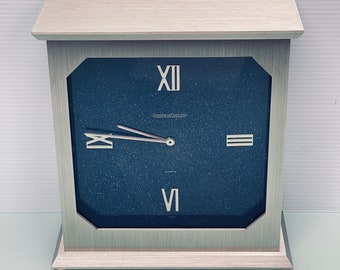 May include: A silver-toned clock with a black face and white Roman numeral markings. The clock face has a subtle sparkle effect. The clock reads "Jardins de la Colline" and "Quartz".