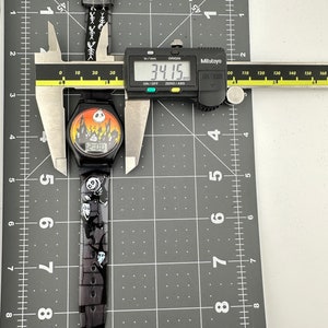 May include: Black wristwatch with a Nightmare Before Christmas design. The watch face features a digital display showing 12:00. The watch band is black with white characters and images of characters from the movie. The watch is measured with a caliper showing 34.15 mm.
