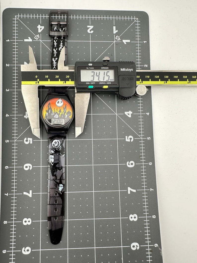 May include: Black wristwatch with a Nightmare Before Christmas design. The watch face features a digital display showing 12:00. The watch band is black with white characters and images of characters from the movie. The watch is measured with a caliper showing 34.15 mm.