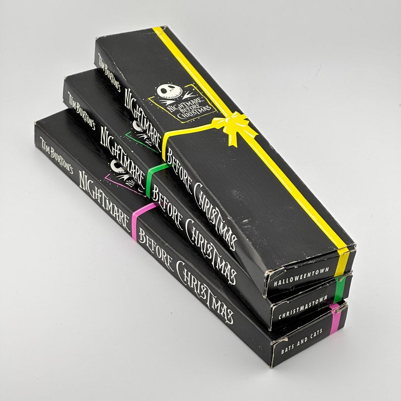 May include: Three black boxes stacked on top of each other. The boxes are wrapped in yellow, green, and pink paper. The boxes are labeled with the text "Tim Burton's Nightmare Before Christmas" and "Halloweentown", "Christmas Town", and "Bats and Cats".