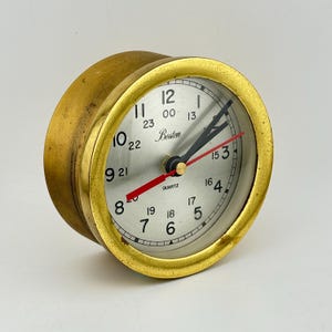 May include: A vintage brass quartz clock with a silver face. The clock face features black numerals, black and red hands, and the word "Boston". The clock has a gold-colored bezel and casing.
