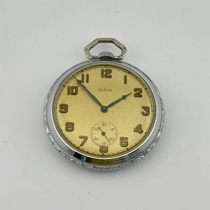 May include: A silver-toned Elgin pocket watch featuring a circular case with an intricate engraved border. The watch face is a muted gold color with black Arabic numerals, dark hands, and a small sub-dial for seconds at the six o'clock position.