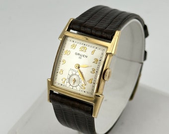 May include: A gold-toned Gruen watch with a white face and black leather band. The watch face has black Roman numeral hour markers and a small seconds dial. The watch is in a square case with rounded corners.