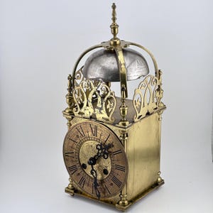 May include: Antique brass clock with a silver bell and ornate detailing. The clock face has Roman numerals and a decorative design.