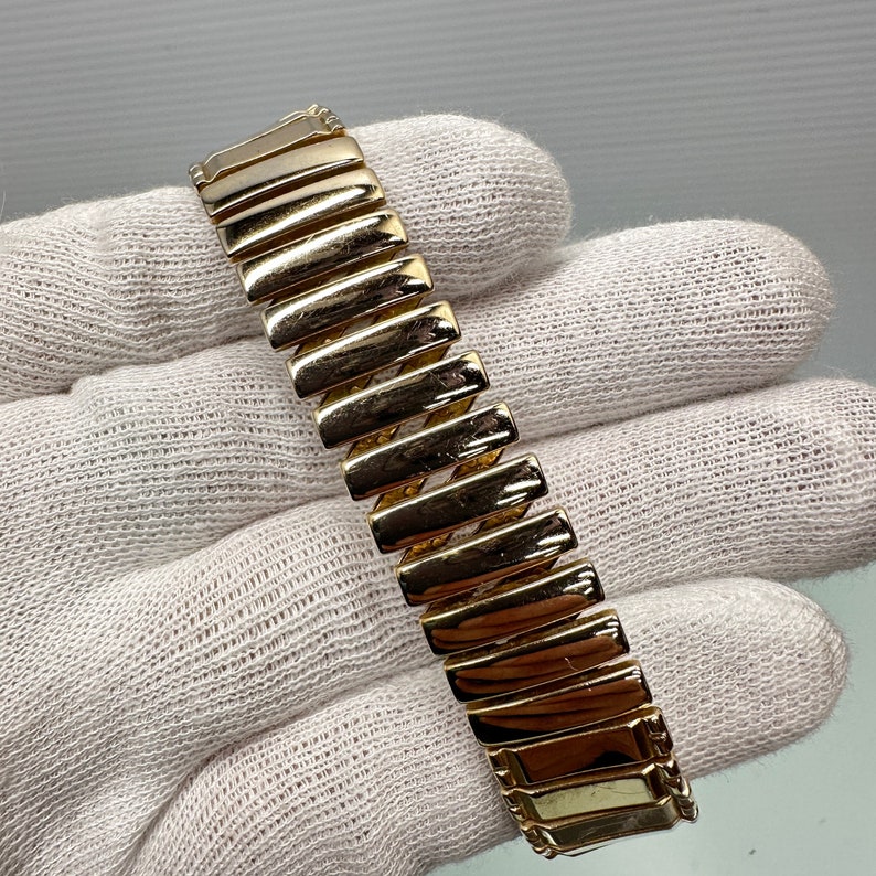 May include: A gold-tone watch band with a textured, ridged design. The band is made of metal and has a polished finish.