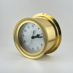 Puede incluir: Un reloj dorado con esfera blanca y manecillas negras. El reloj tiene forma redonda con un bisel dorado y un diseño circular. La esfera muestra los números del 1 al 12.