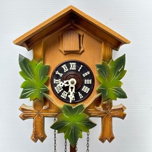 May include: A wooden cuckoo clock with a light brown frame, a black clock face with white Roman numerals, and green leaf accents. The clock has decorative wooden crosses and hanging chains.