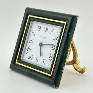 May include: A square Cartier desk clock with a green frame and gold trim. The clock face is white with black Roman numerals and hands. The clock is supported by a gold-colored stand.