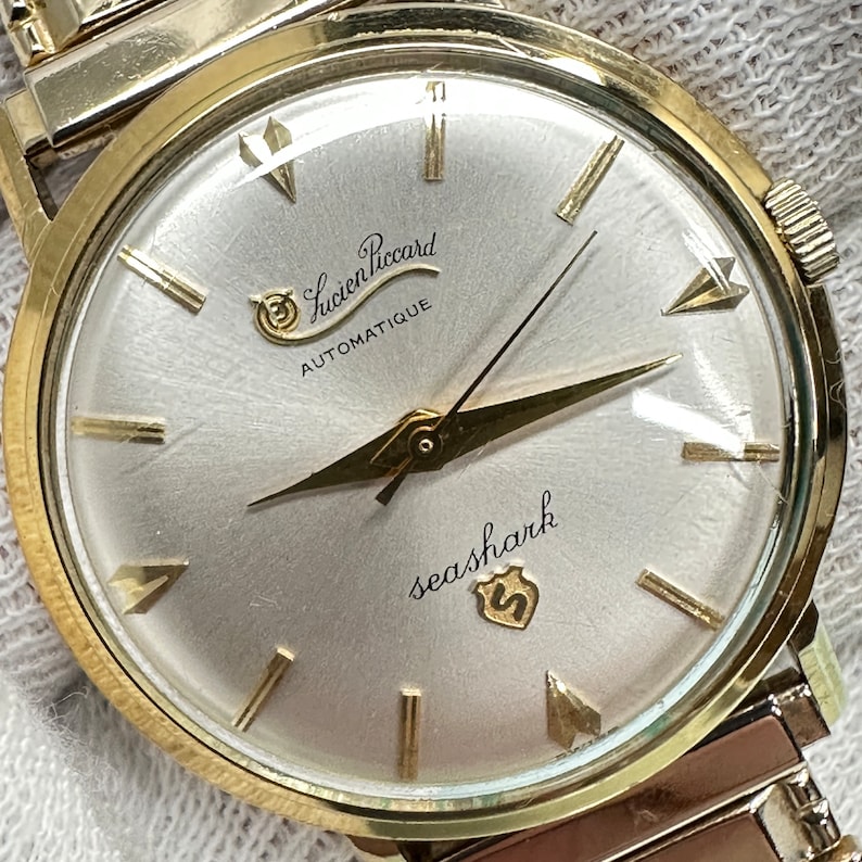 May include: A gold-tone wristwatch with a white face. The watch face has black Roman numeral hour markers and a black second hand. The watch face also has the text "Lucien Piccard Automatique" and "seashark" with a shield logo.