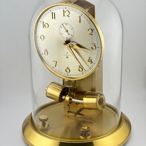 May include: A gold-toned clock with a cream-colored face, protected by a clear glass dome. The clock face features black numerals and hands, with a smaller dial at the top. The base and other components are also gold-toned.