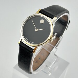 May include: A black-faced wristwatch with a silver and gold band and a black leather strap. The watch face has a small white circle at the 12 o'clock position and the words "MOVADO" and "ZENITH" printed on the face.
