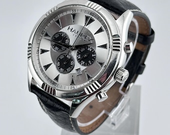 May include: A silver chronograph wristwatch with a black leather band. The watch face has a white background with black markings and a date window at the 4 o'clock position. The watch face also has the text "HAUREX" printed on it.
