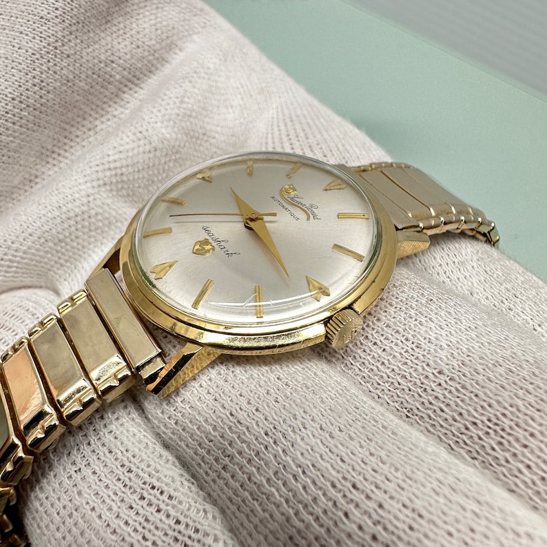 May include: A gold-toned wristwatch with a white face and black hands. The watch has a gold-toned metal band and says "Sea-Land" and "Automatic" on the face.