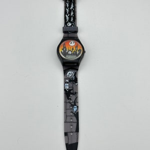 May include: Black digital wristwatch with a Nightmare Before Christmas theme. The watch face features a scene from the movie with Jack Skellington's silhouette and the words "This is Halloween". The watch band is black with a repeating pattern of Jack Skellington's head and the words "This is Halloween".