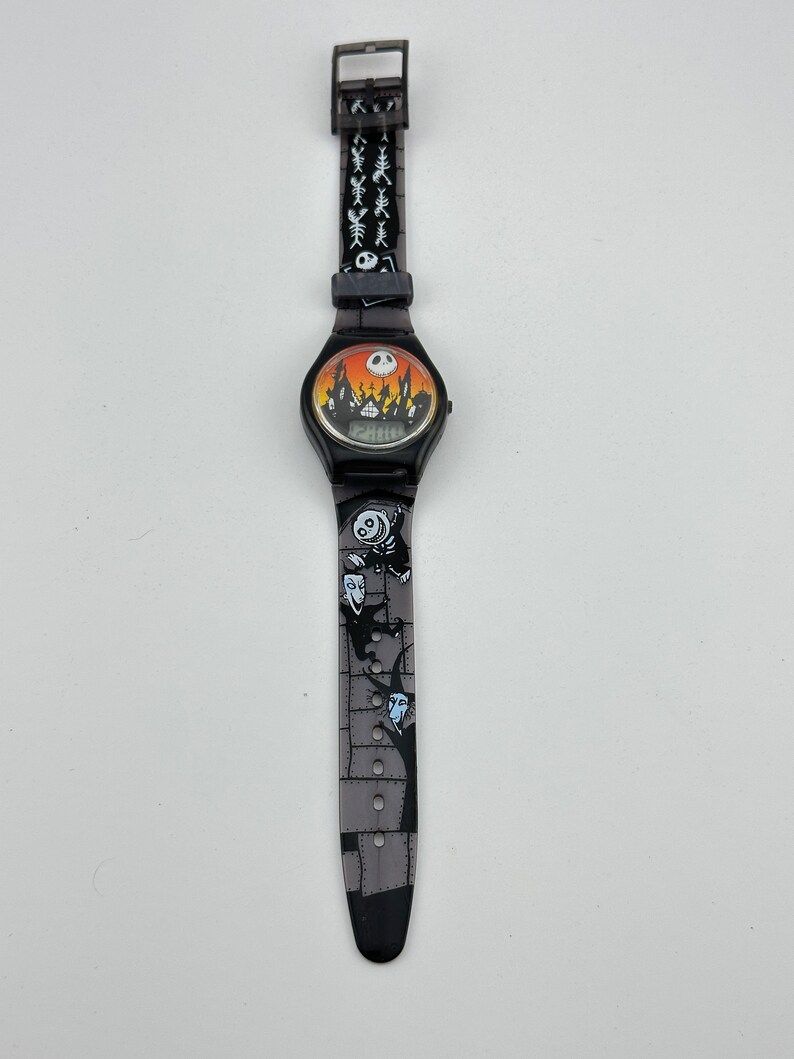 May include: Black digital wristwatch with a Nightmare Before Christmas theme. The watch face features a scene from the movie with Jack Skellington's silhouette and the words "This is Halloween". The watch band is black with a repeating pattern of Jack Skellington's head and the words "This is Halloween".