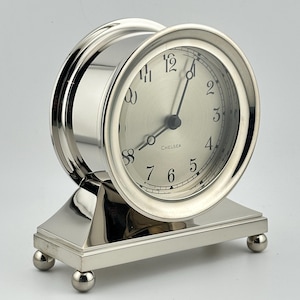 May include: A silver-toned metal desk clock with a polished finish, featuring a round face with black Arabic numerals and the word Chelsea printed in the center. The clock is mounted on a rectangular base supported by four small spherical feet.