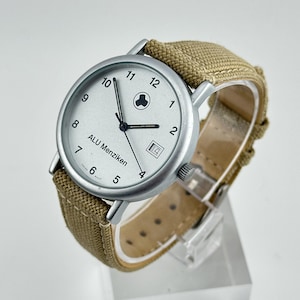 May include: A silver-toned wristwatch with a white face, black hands, and a date window at the 3 o'clock position. The watch features the text "ALU Menziken" and a canvas band in a tan color. The watch is displayed on a clear stand.