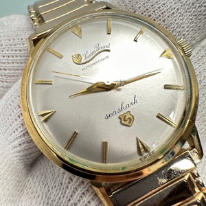 May include: A gold-tone wristwatch with a white face and black hands. The watch face has the text "Lucien Piccard Automatique" and "seashark" with a small logo. The watch has a gold-tone metal band.