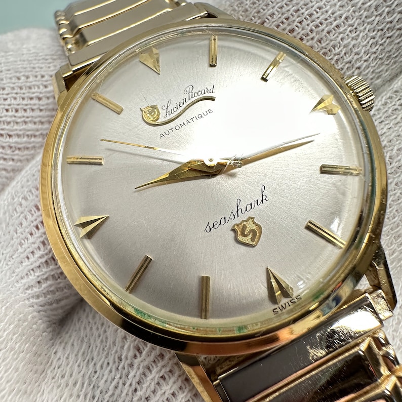 May include: A gold-tone wristwatch with a white face and black hands. The watch face has the text "Lucien Piccard Automatique" and "seashark" with a small logo. The watch has a gold-tone metal band.