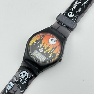 May include: Black and gray digital watch with a Nightmare Before Christmas design. The watch face features a black silhouette of the Haunted Mansion against a sunset background. The watch band has a repeating pattern of skeletons and bones.