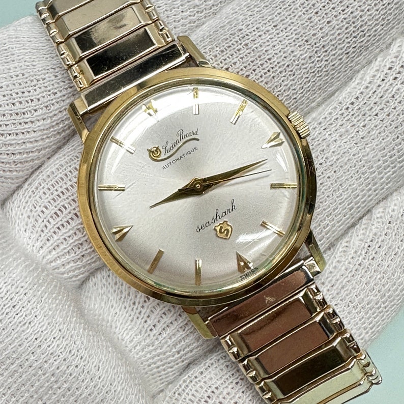 May include: A gold-tone wristwatch with a white face and black hands. The watch has a gold-tone band and says "Leon Picard Automatique" and "seashark" on the face.