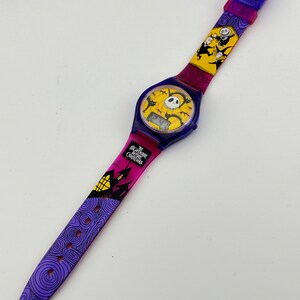 May include: A purple, yellow, and black wristwatch with a digital display and a band featuring characters from The Nightmare Before Christmas. The band has a purple, yellow, and pink gradient.