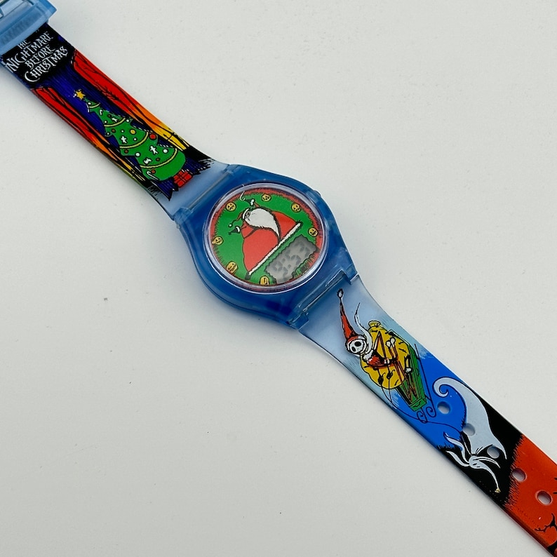 May include: A blue and orange wristwatch with a digital display featuring a Santa Claus character. The watch band has a colorful design with images from the movie "The Nightmare Before Christmas".