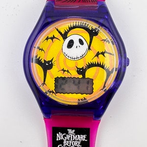 May include: A pink and blue digital watch with a yellow face featuring Jack Skellington from The Nightmare Before Christmas. The watch face also has bats and cats flying around Jack Skellington's face. The watch band has the words "The Nightmare Before Christmas" printed on it.