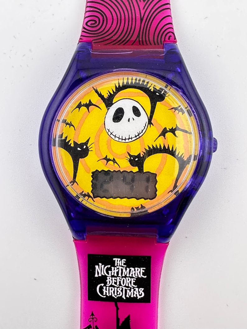 May include: A pink and blue digital watch with a yellow face featuring Jack Skellington from The Nightmare Before Christmas. The watch face also has bats and cats flying around Jack Skellington's face. The watch band has the words "The Nightmare Before Christmas" printed on it.
