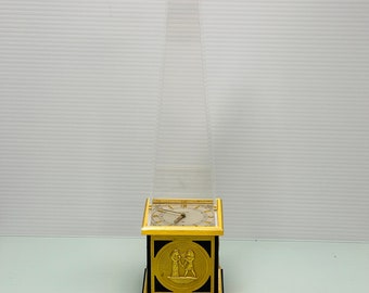 May include: A gold-toned clock with an Egyptian-inspired design. The clock is in the shape of a square with a clear glass pyramid on top. The clock face is white with black Roman numeral hour markers and black hands. The clock is sitting on a black surface.