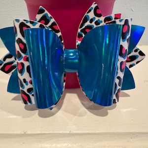 Puede incluir: Un lazo para el cabello hecho a mano con un llamativo acabado azul iridiscente. El diseño del lazo incorpora un llamativo estampado de leopardo con manchas negras, rosas y azules, creando un accesorio llamativo y juguetón.