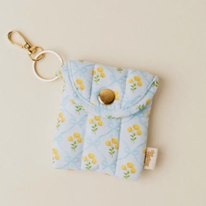 以下が含まれることがあります： 黄色い花の模様がついた小さな青いキルティングポーチ。金色のジッパーと金色のボタン留め具が付いています。金色のキーリングも付いています。