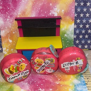 May include: Three round pink containers featuring Mini Verse Make It Mini Plush branding, each with a handwritten white label reading Emmett Pic, Shelby Pic, and Dipper B, positioned in front of a small pink and yellow toy display shelf.