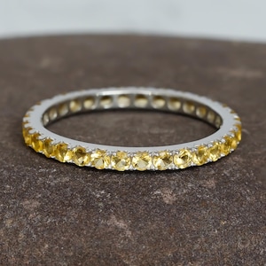 Peut inclure: Une délicate bague éternelle avec une rangée de petites pierres jaunes rondes serties dans une bande argentée.