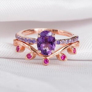 May include: A rose gold ring set featuring an oval-cut amethyst center stone. The band is adorned with small purple and pink gemstones. The ring sits on a white, textured surface.