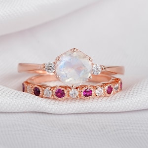 May include: A set of two rose gold rings with a large moonstone center stone and smaller diamonds on the band. The bottom ring has a row of small diamonds and pink gemstones.