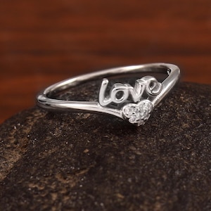 May include: A silver ring spelling out the word "love" in cursive. The ring has a small heart shape with clear stones. The ring is displayed on a dark, textured surface.