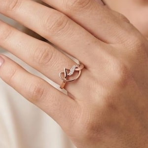 May include: Rose gold ring with a delicate heart-shaped design, adorned with small gemstones. The ring is displayed on a hand, highlighting its elegant and minimalist aesthetic. A piece of jewellery suitable for daily wear or special events.