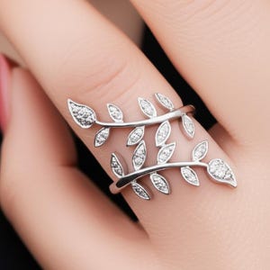 May include: A silver-colored ring with a leaf design. The ring features two branches of leaves, each with small, sparkling stones. The leaves are arranged to wrap around the finger, creating a nature-inspired accessory.