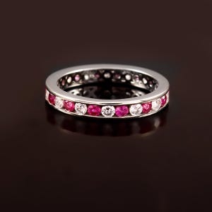 May include: A silver eternity ring featuring alternating round-cut rubies and clear gemstones. The gemstones are set in a continuous band around the ring. The ring is displayed on a dark reflective surface.