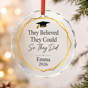 May include: A circular clear glass ornament with a faceted edge, featuring a gold geometric frame, a black graduation cap icon, and the text "They Believed They Could So They Did" followed by the name "Emma" and the year "2026" in black ink.