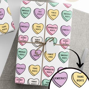 May include: White wrapping paper with a repeating pattern of pastel-colored heart-shaped candies. Each candy has a different phrase, including "Trans Rights," "Immigrant Rights," and "Democracy." The wrapped gifts are tied with twine.