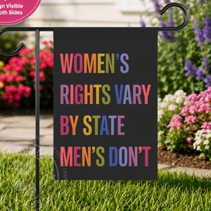 Może przedstawiać: Prostokątna czarna flaga ogrodowa z wielokolorowym napisem "WOMEN'S RIGHTS VARY BY STATE MEN'S DON'T" wykonanym pogrubioną czcionką bezszeryfową. Flaga jest zawieszona na czarnym metalowym stojaku i zadrukowana po obu stronach.