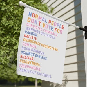 May include: A white flag displaying the text "NORMAL PEOPLE DON'T VOTE FOR" and a list of groups in different colours. The flag is mounted on a white pole with a black bracket, seen outside.