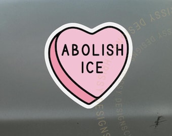 May include: A pink heart-shaped sticker with the words "ABOLISH ICE" in black capital letters. The sticker has a white border and is placed on a grey surface.