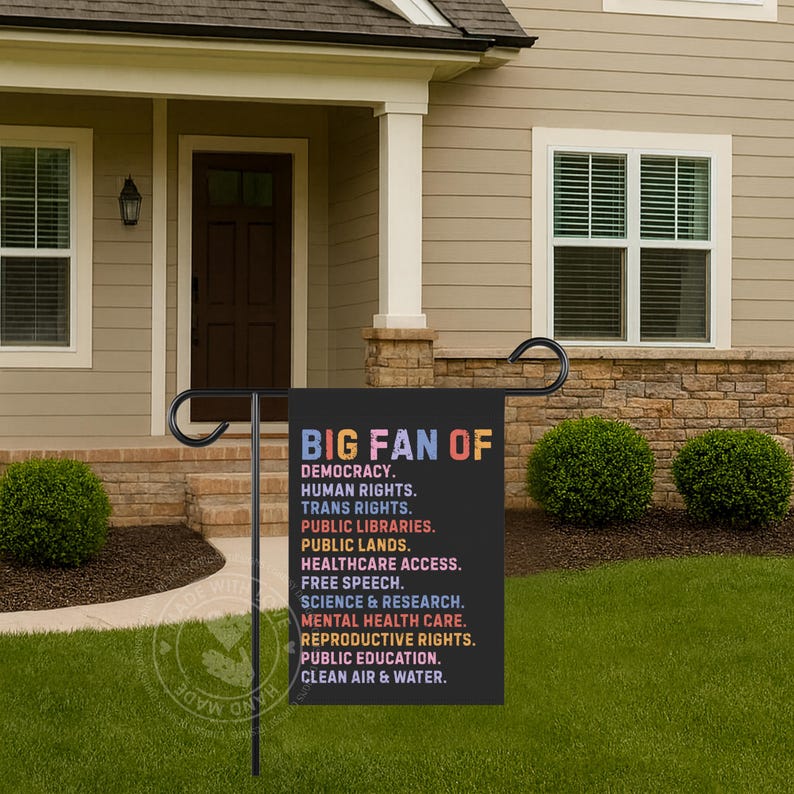 May include: A black garden flag with the text "BIG FAN OF" in colorful letters. Below, a list of values including "DEMOCRACY," "HUMAN RIGHTS," and "CLEAN AIR & WATER" is displayed in white text. The flag is displayed in a front yard.