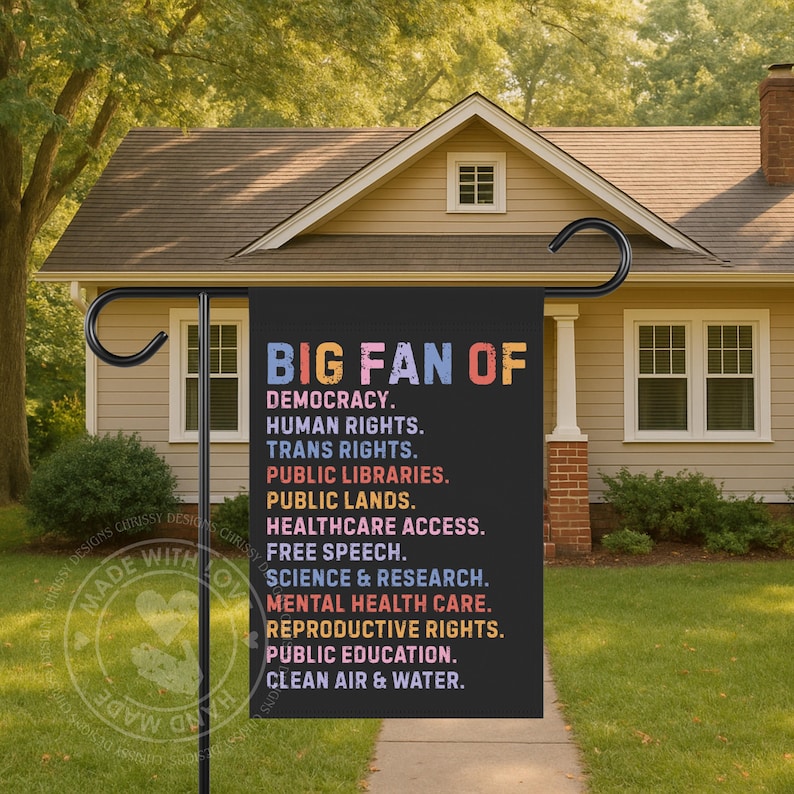 May include: A black garden flag with the text "BIG FAN OF" in colorful letters. Below, a list of values including democracy, human rights, and healthcare access is displayed. The flag is hanging in front of a house.