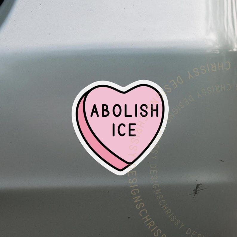Puede incluir: Una pegatina rosa en forma de coraz&oacute;n con las palabras "ABOLISH ICE" en letras may&uacute;sculas negras. La pegatina tiene un borde blanco y est&aacute; colocada sobre una superficie gris.